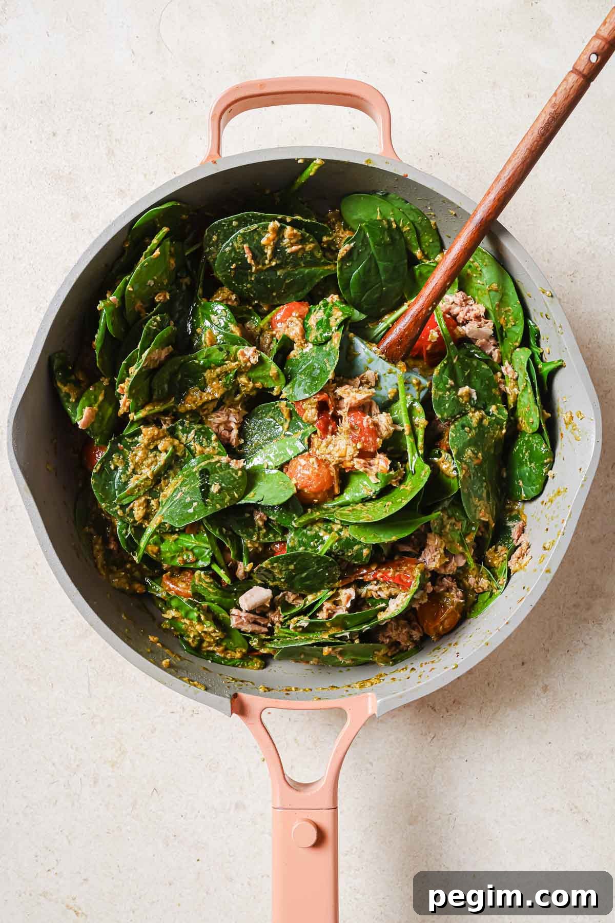 A skillet filled with spinach, tomatoes, and tuna with a wooden spoon.
