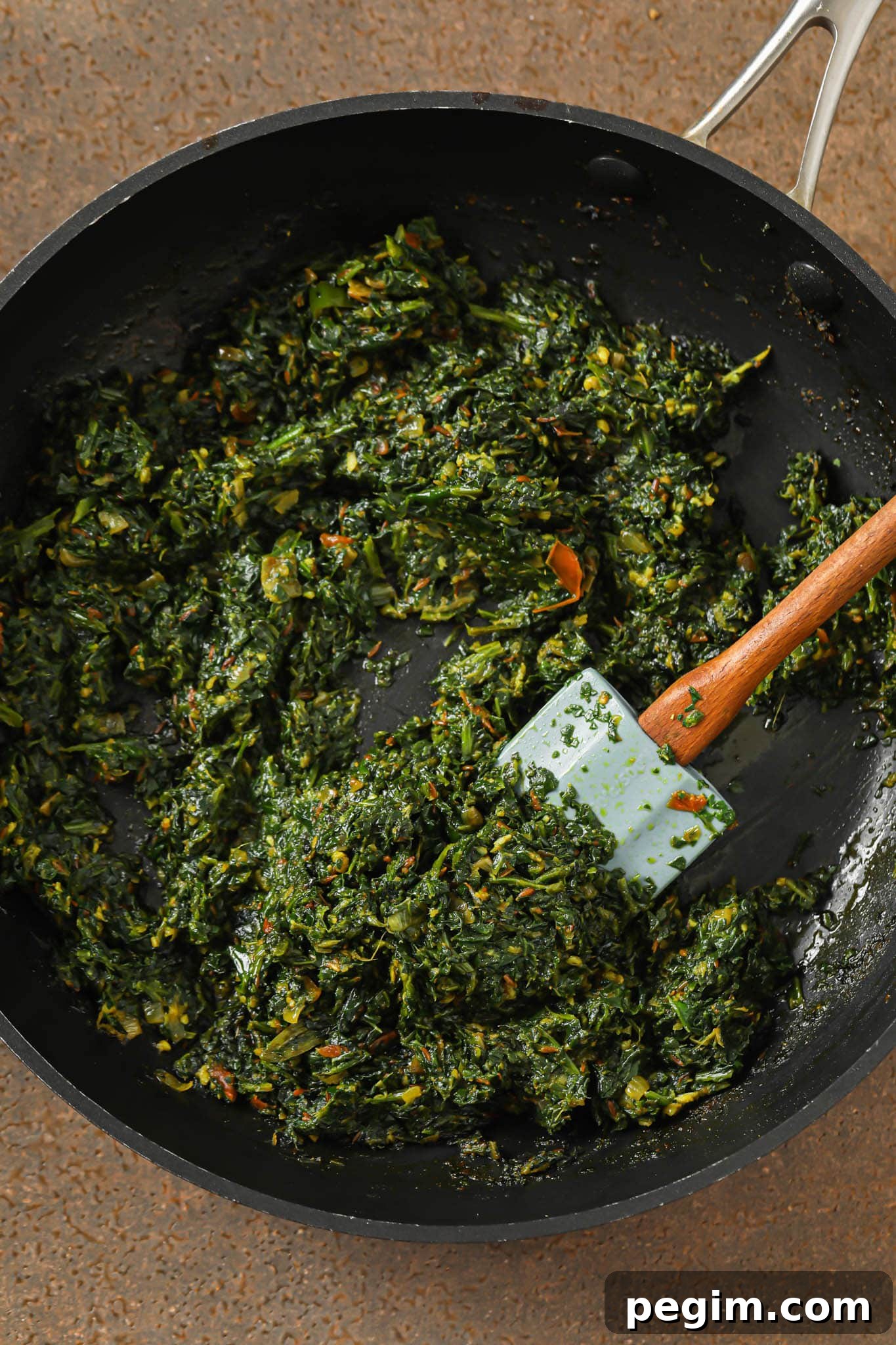 Cooked down spinach-onion-tomato mixture in a skillet with a blue spatula.