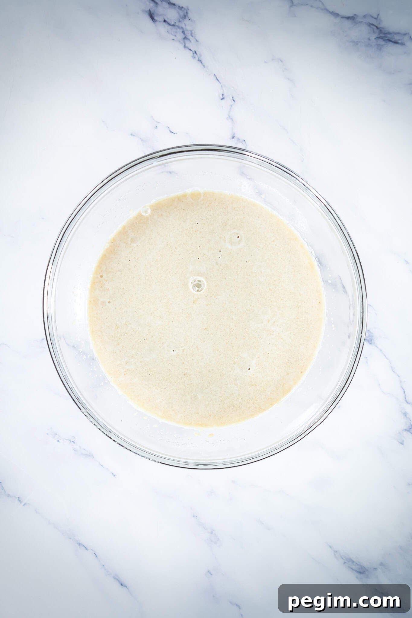 A glass bowl filled with light beige batter sits on a white marble surface