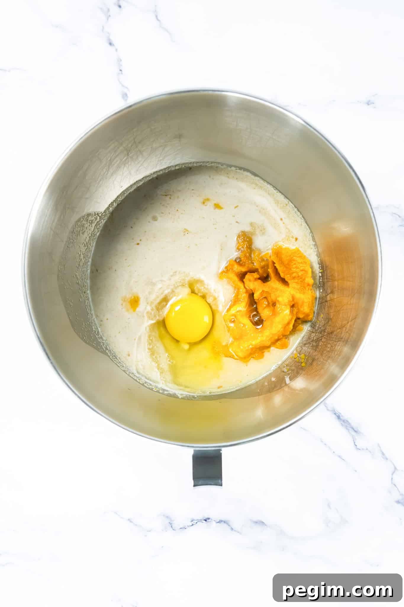 A metal mixing bowl with an egg, pumpkin puree, and liquid ingredients on a white marble surface