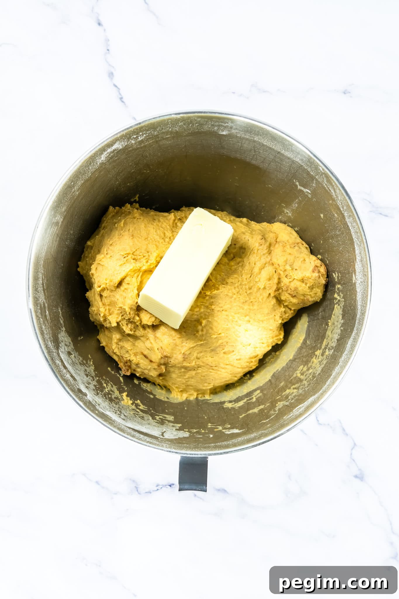 A metal mixing bowl with yellow dough and a stick of butter on top, seen from above on a light surface
