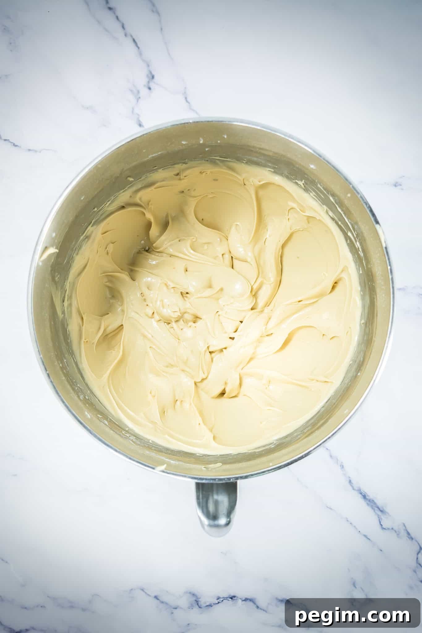 A metal mixing bowl filled with smooth, creamy frosting for Pumpkin Cinnamon Rolls rests on a white marble surface.
