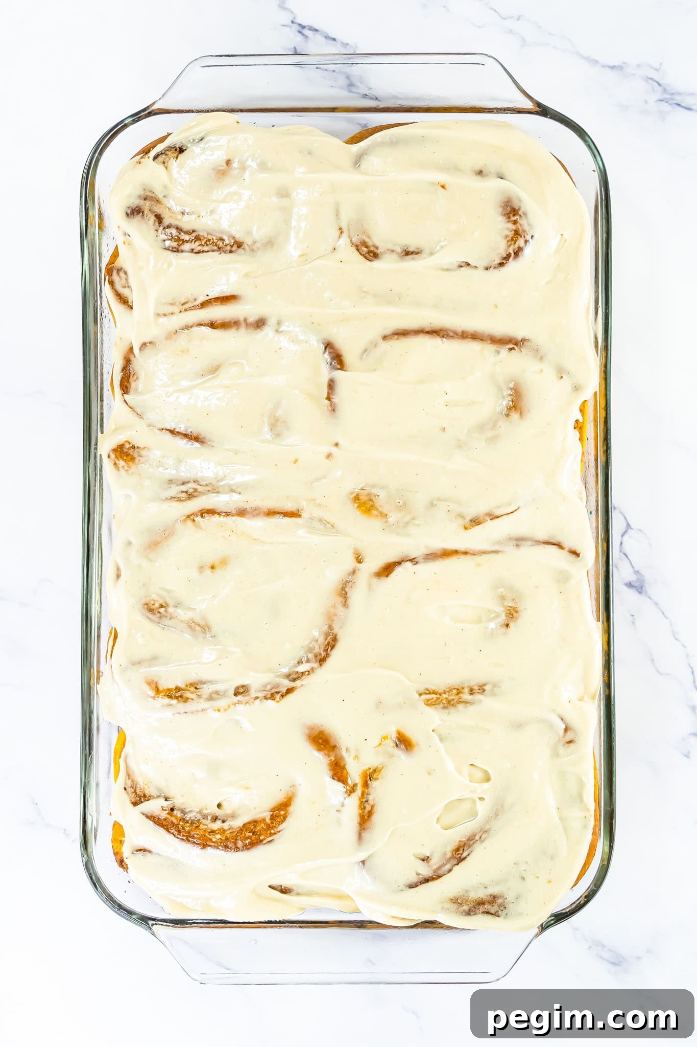 A glass baking dish with frosted Pumpkin Cinnamon Rolls sits on a white marble surface.