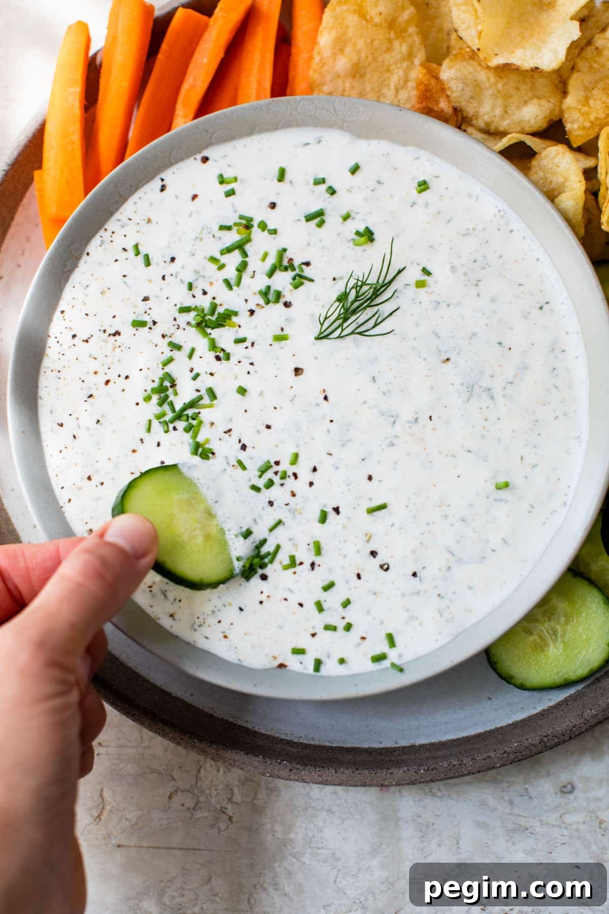 someone's hand dipping a cucumber into a bowl of white-colored dip