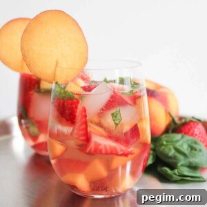 Two glasses of sparkling rose sangria garnished with thin peach slices in front of a display of summer fruits.
