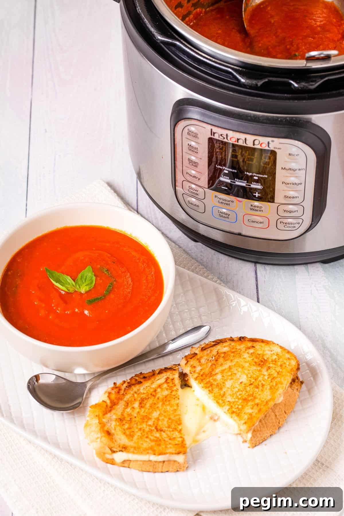 Close up shot of finished tomato soup in a white bowl, with a grilled cheese toastie on a white plate, instant pot in the background.