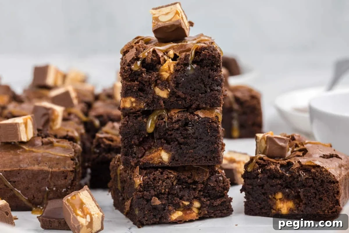 A stack of snickers brownies with caramel and candy pieces, surrounded by more brownies and candy chunks on a white surface.