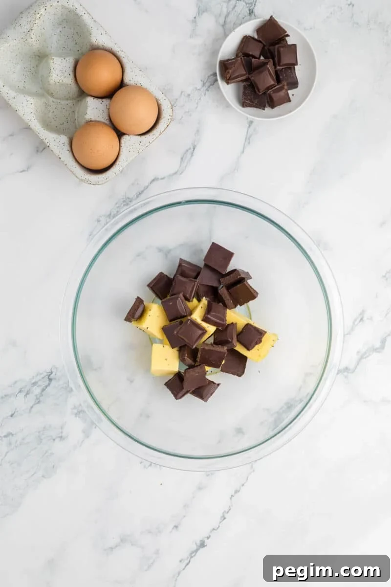 A glass bowl with chunks of butter and dark chocolate, next to a dish of eggs and a small plate of chocolate pieces on a marble countertop.