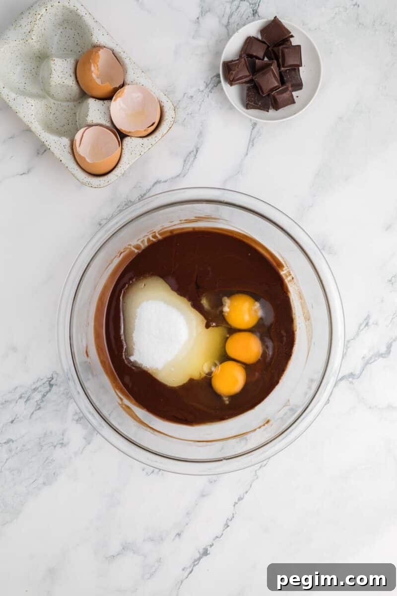 A glass bowl with chocolate mixture, sugar, and three eggs sits on a marble counter beside cracked eggshells and a small bowl of chocolate pieces.