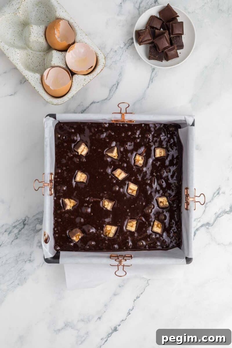 A square pan lined with parchment paper filled with chocolate brownie batter and peanut butter chunks, next to a bowl of egg shells and a dish of dark chocolate pieces on a marble surface.