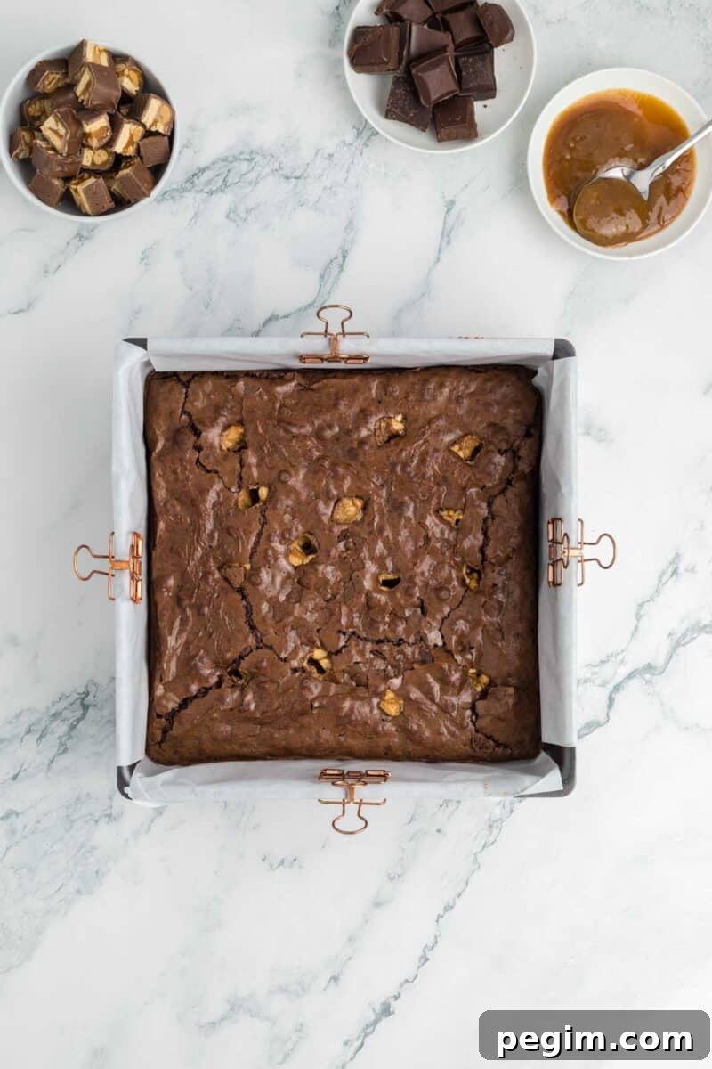 A pan of snickers brownies with caramel pieces is on a marble surface, surrounded by bowls of caramel sauce, chopped chocolate, and caramel candies.
