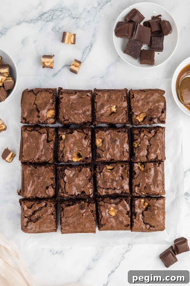 A batch of brownies cut into sixteen squares on parchment paper, with bowls of chocolate pieces and caramel sauce nearby on a marble surface.