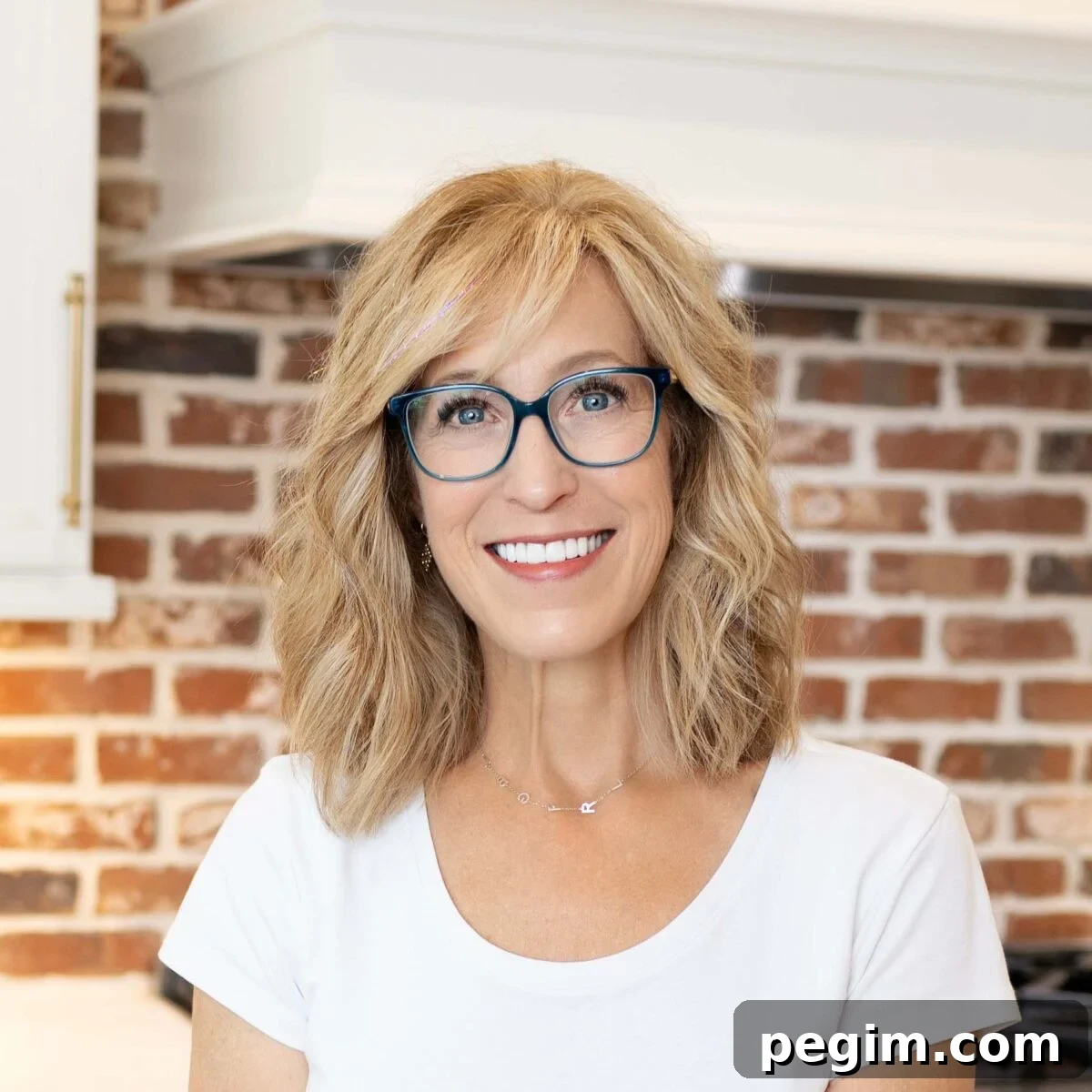 Person with shoulder-length blond hair and blue glasses smiling indoors in front of a red brick wall.