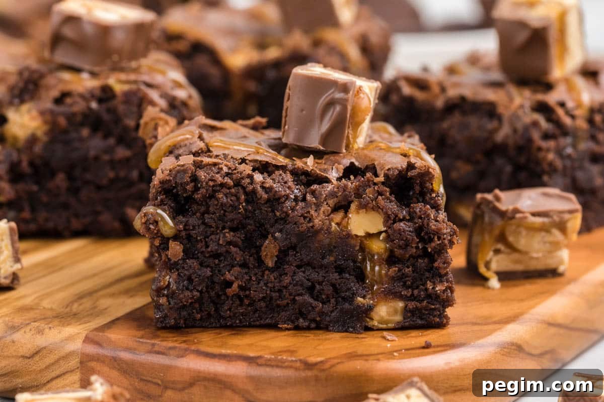A close-up of a chocolate brownie with snickers with caramel and nut pieces, topped with a small chocolate candy, on a wooden board.