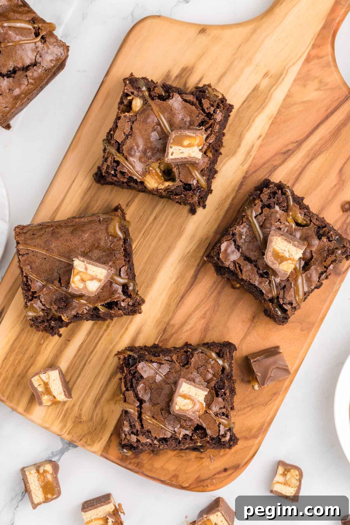 Four snickers brownies topped with pieces of candy bars are arranged on a wooden board, with additional candy pieces scattered around on a white surface.