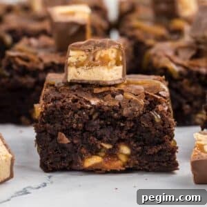 A close-up of a snickers brownie with chopped nuts, topped with a piece of caramel and nougat candy, with more brownies in the background.