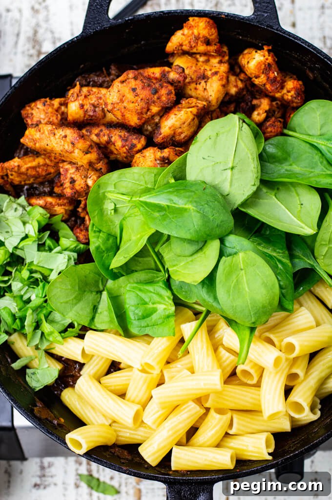 A cast iron skillet with chicken, rigatoni pasta noodles, fresh spinach and fresh basil.