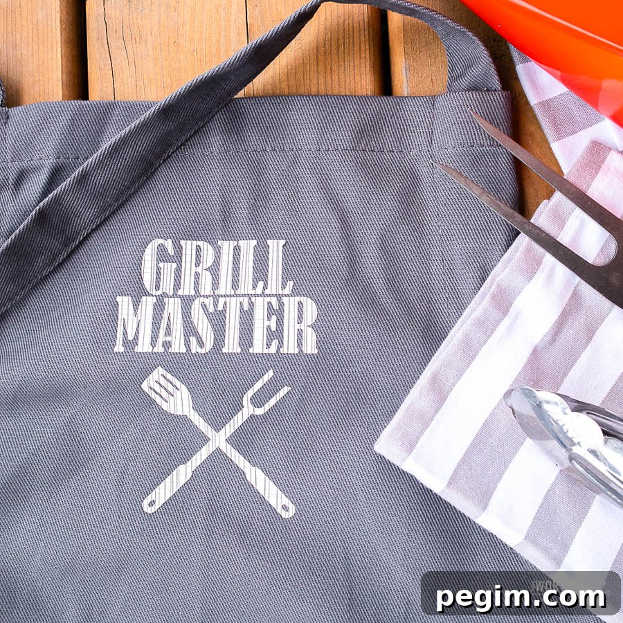 Grey DIY apron with white 'grill master' message on front laying on table by grill utensils