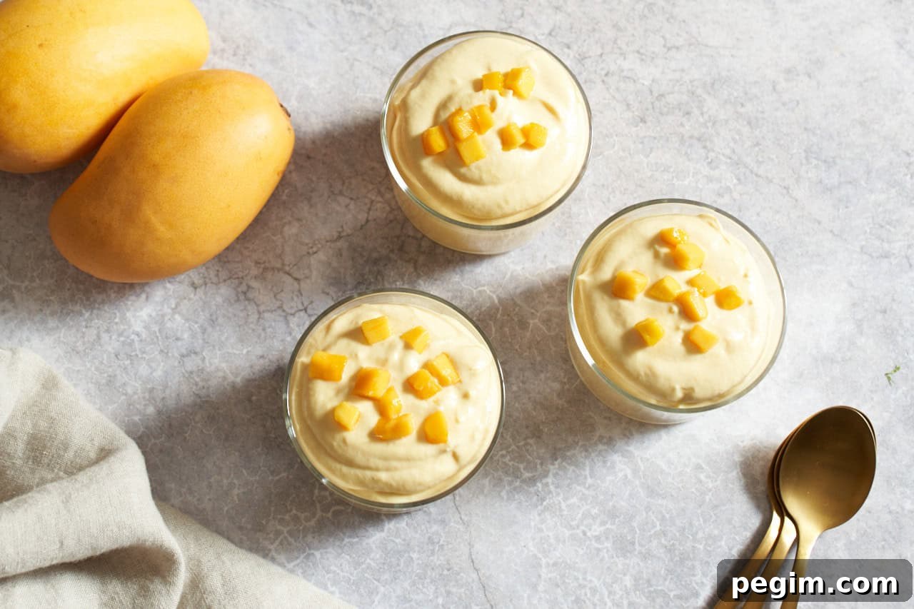 Three servings of mango mousse in small glasses topped with diced fresh mango, three gold spoons are on the right, two yellow mangoes are on the left.