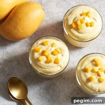 Three servings of mango mousse in small glasses topped with diced fresh mango, three gold spoons are on the right, two yellow mangos are on the left.