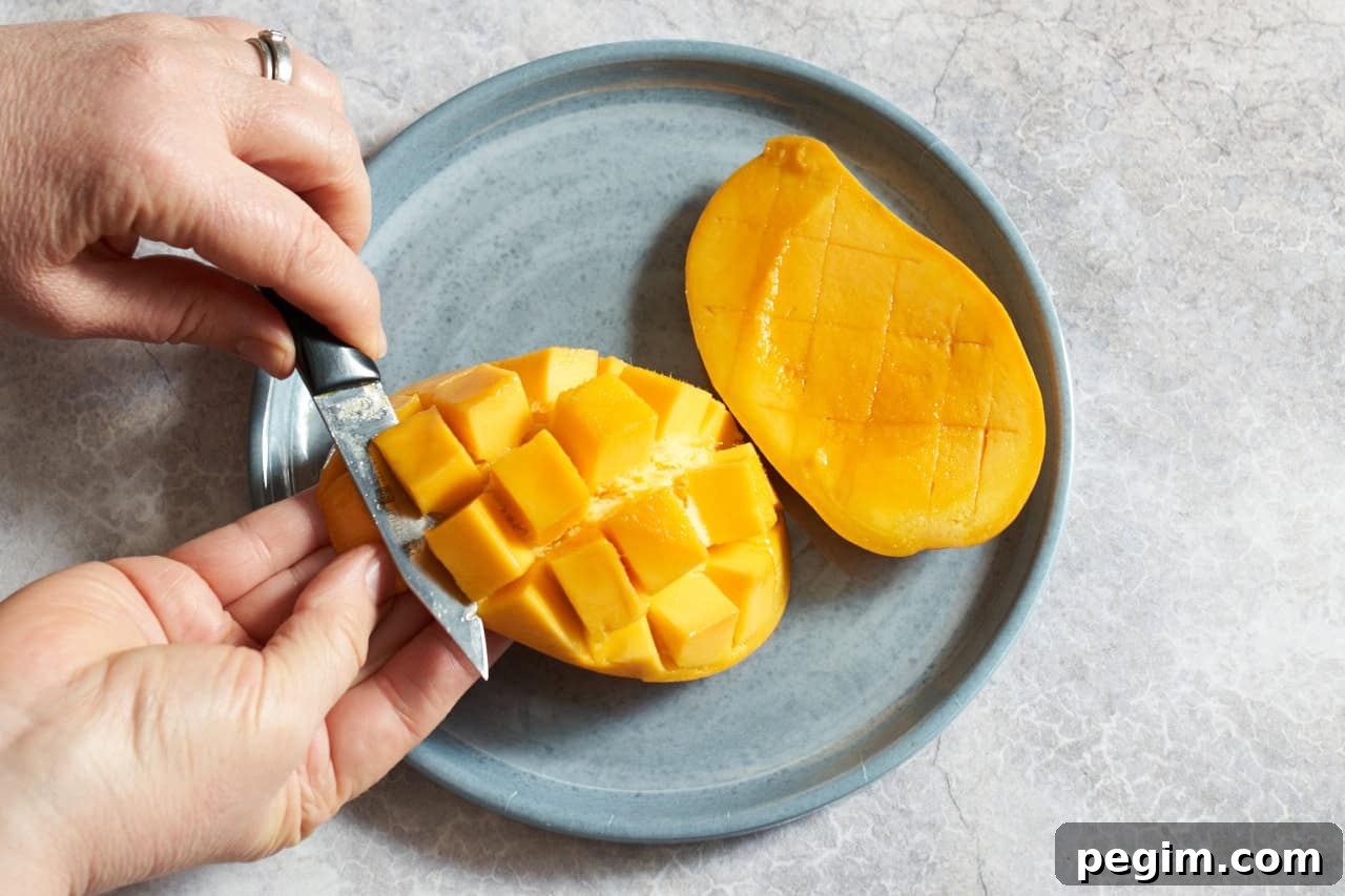 A woman's hands shown cutting a mango that has been cross-hatched into chunks away from the peel, another mango half is on the right.