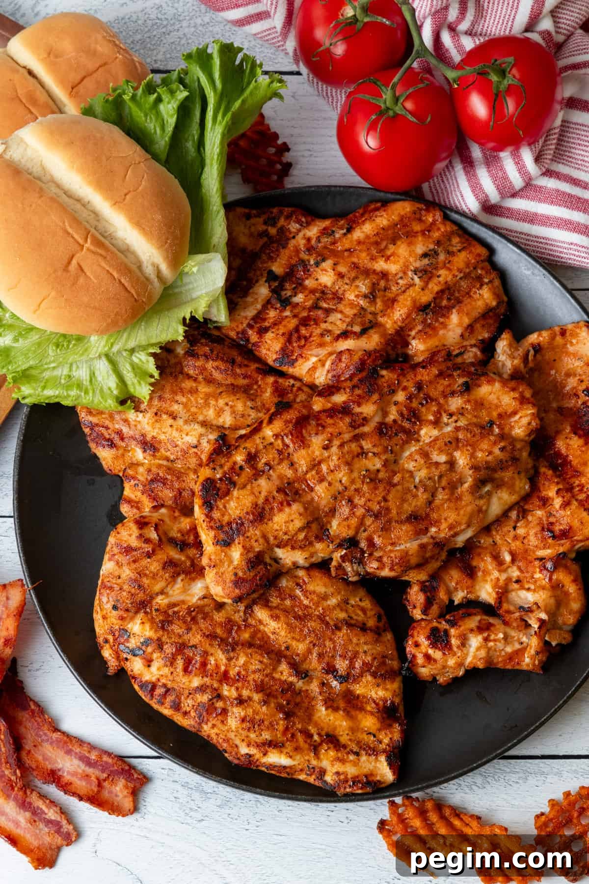 Grilled chicken breasts on black plate with buns, lettuce, and tomatoes around plate.