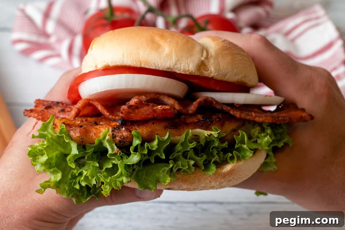 Chicken sandwich with lettuce, onion, tomatoes, and bacon being held with two hands.