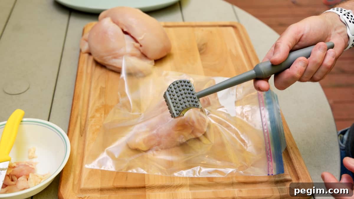 Chicken breasts in ziplock bag on cutting board with a meat mallet being held by hand over chicken.