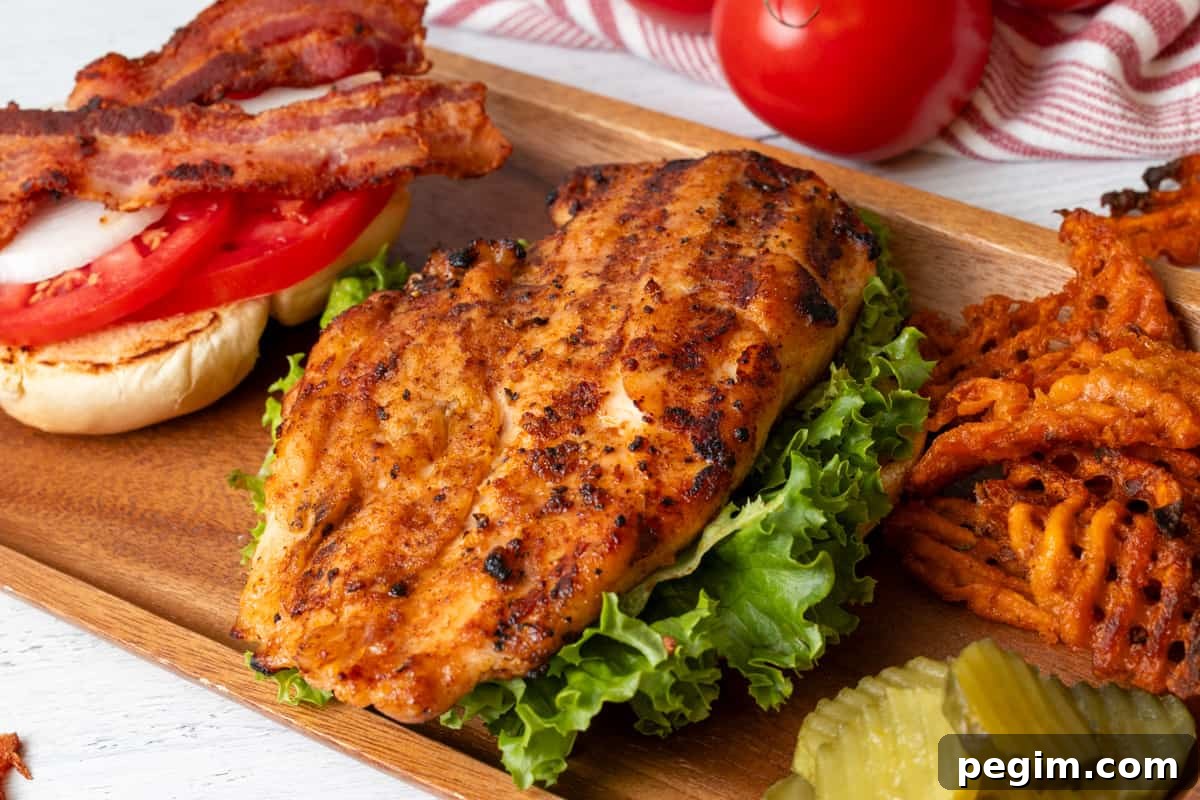 Grilled Chicken breast on lettuce with a bun next to it with onion, tomato, and bacon.