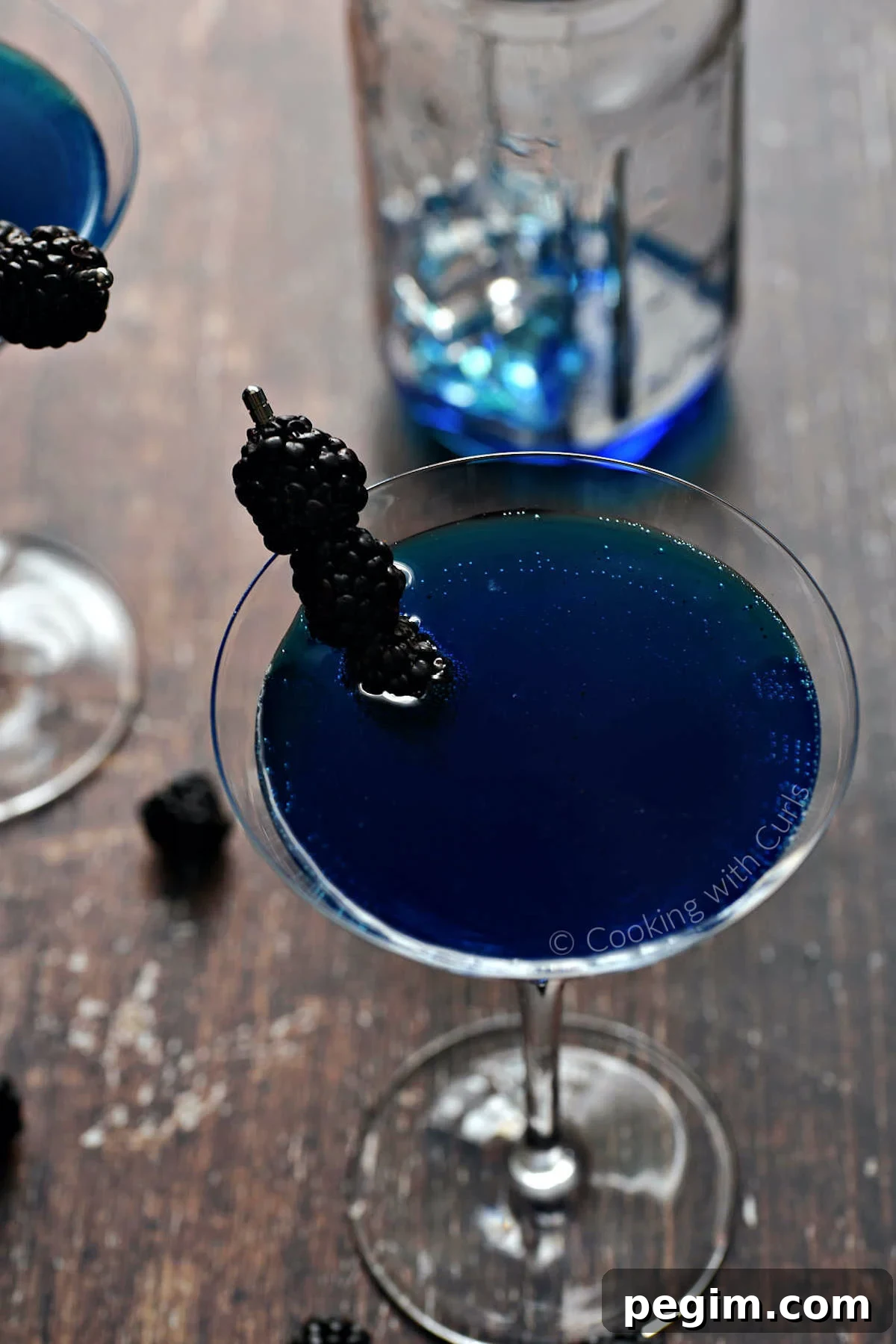A martini glass filled with a dark royal blue martini that's garnished with three blackberries on a skewer.