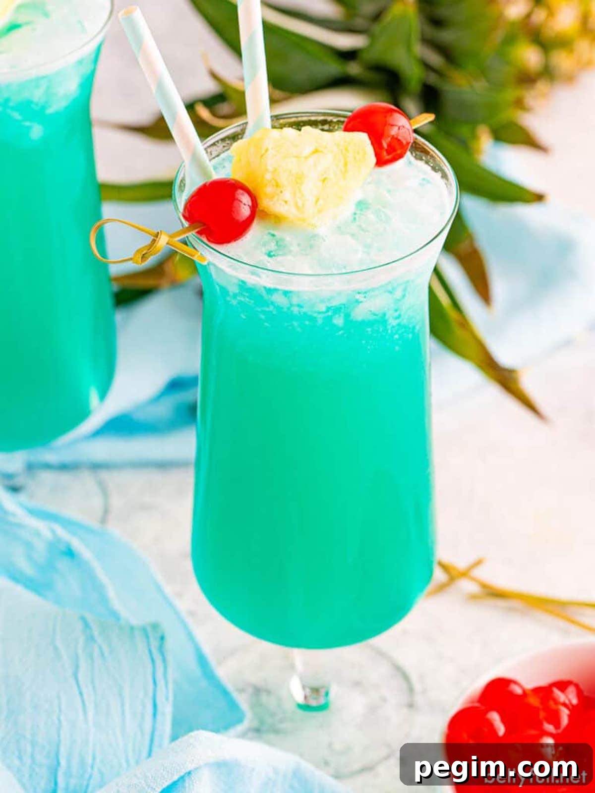 A tall hurricane glass filled with a bright blue Blue Hawaiian cocktail that's garnished with two cherries and a pineapple wedge on a skewer.
