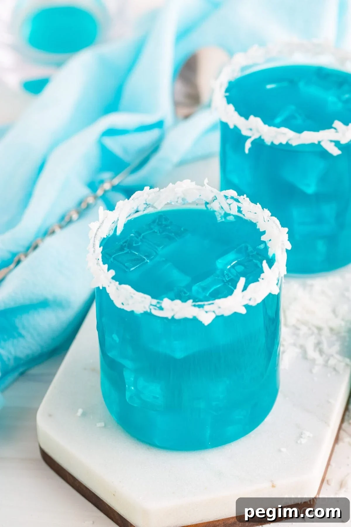 Two old fashioned glasses filled with a bright blue Ocean water cocktail. Each has a rim of shredded coconut.