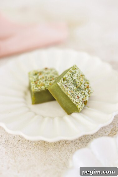 two squares of cake topped with pistachio and coconut on a white plate.