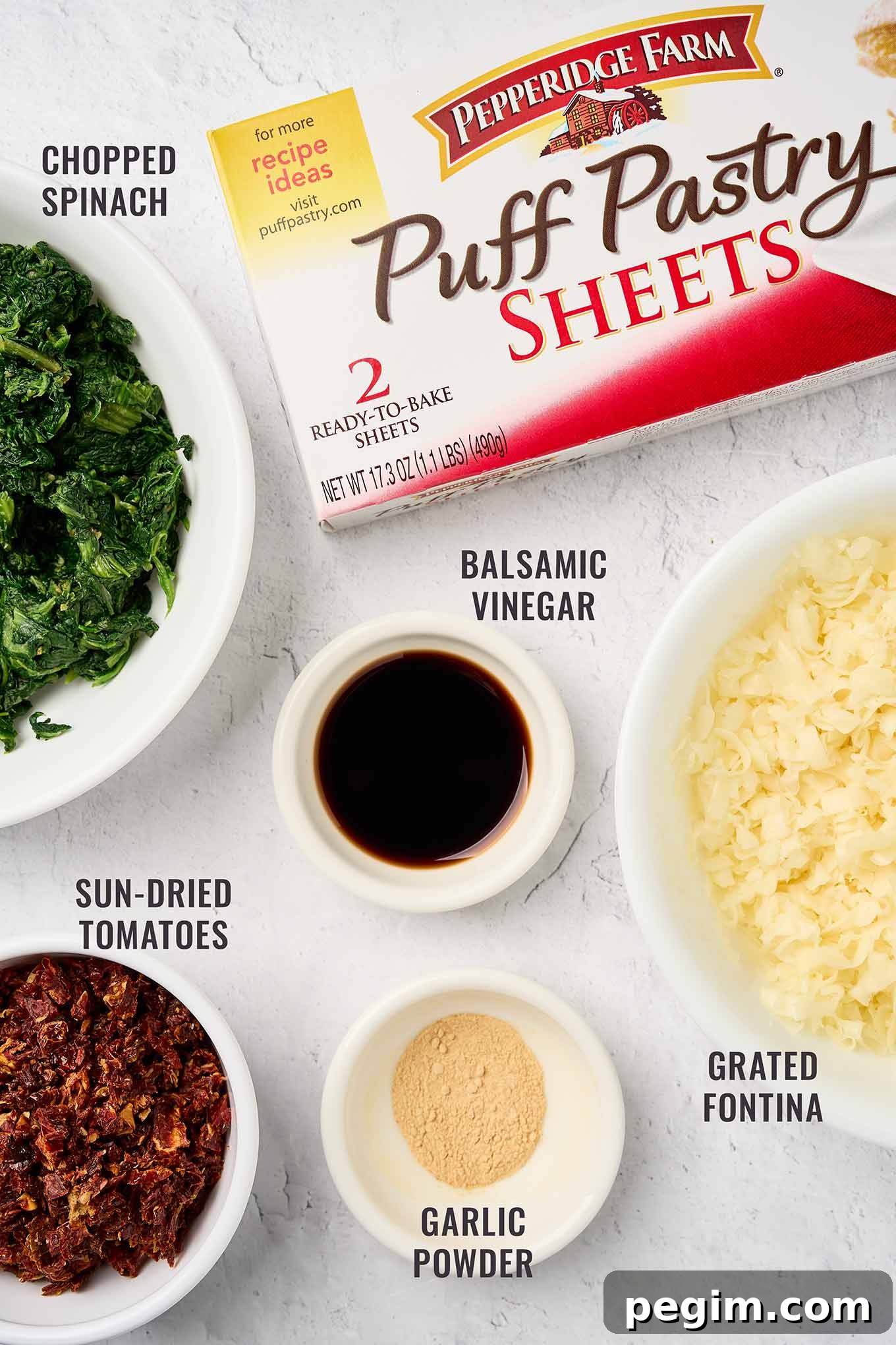 A labeled view of the ingredients for spinach pinwheels, including puff pastry, chopped spinach, sun-dried tomatoes, grated fontina cheese, balsamic vinegar, and garlic powder.