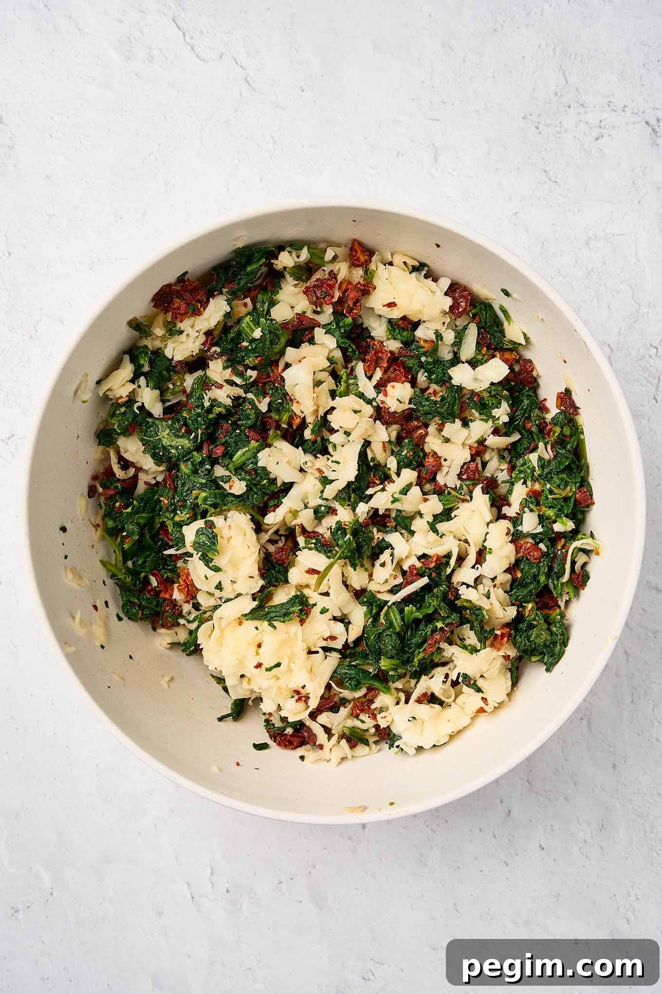 Spinach pinwheel filling mixture in a bowl ready for mixing; chopped spinach, sun-dried tomatoes, and grated fontina.