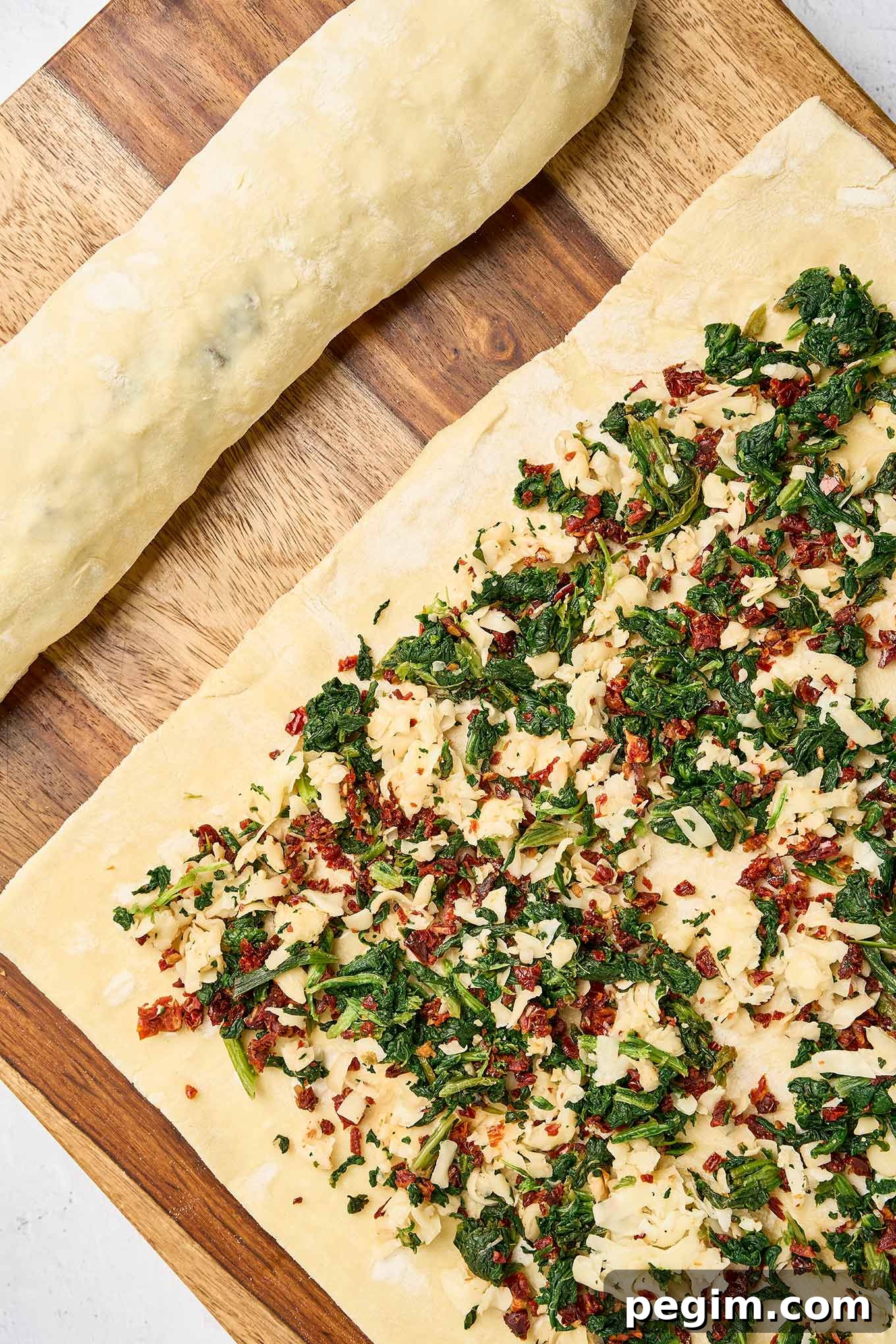 Puff pastry sheet topped with spinach, sun-dried tomatoes, and cheese filling, shown next to an already rolled spinach pinwheel log.