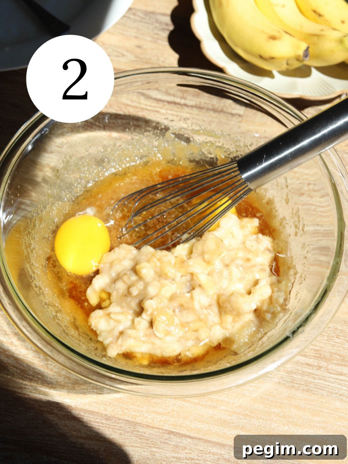A glass mixing bowl with eggs, mashed banana and sugar and butter being whisked together.