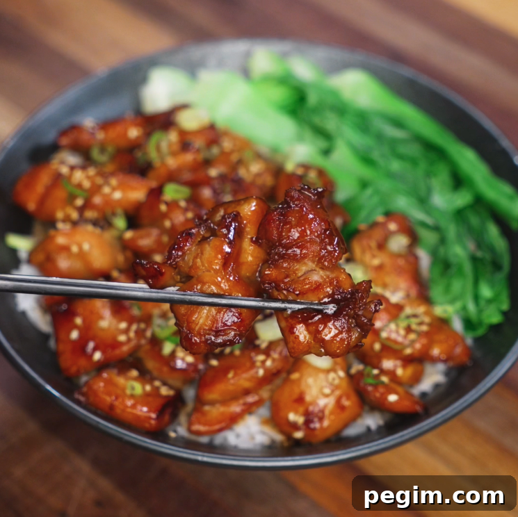 A glossy bite of soy butter chicken is lifted with chopsticks from a bowl filled with sesame-topped chicken, steamed rice, and sautéed bok choy, showcasing its sticky texture and caramelized exterior.