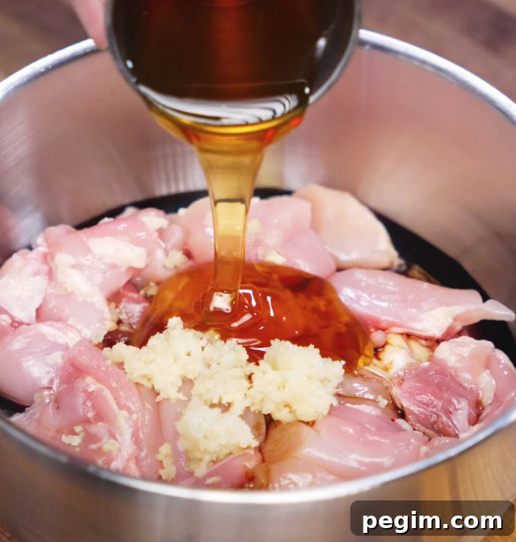A stream of golden honey is poured over a bowl of raw chicken thighs mixed with minced garlic and soy sauce, highlighting the beginning of the soy butter chicken marinade.