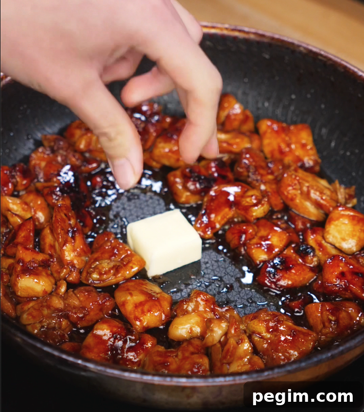 A cube of butter is added to a skillet of sizzling soy-glazed chicken, ready to melt and enrich the sauce with a silky, savory finish for the soy butter chicken.