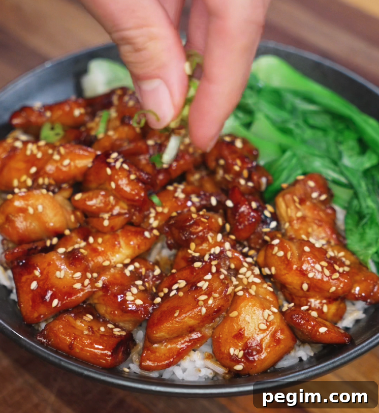 A hand sprinkles chopped scallions over a bowl of soy butter chicken glazed with sesame seeds, served over white rice with bok choy — the final garnish for color, crunch, and freshness.