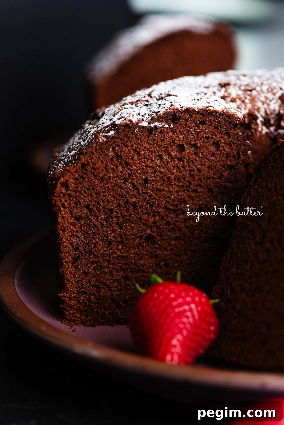 Chocolate chiffon cake sliced with whole strawberries placed on and in front of a dark purple ceramic plate on a black background.