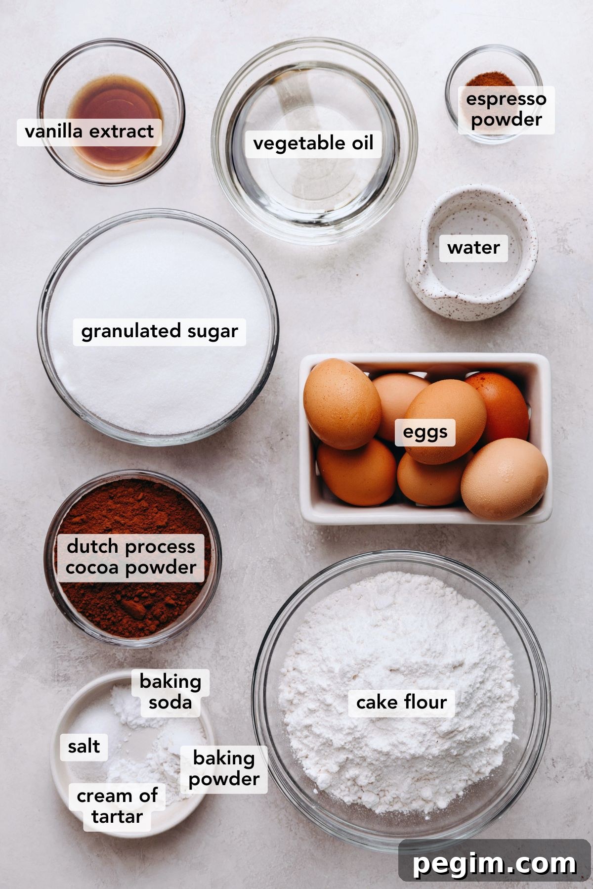 Ingredients in various size plates and bowls labeled as vanilla extract, vegetable oil, espresso powder, granulated sugar, water, eggs, dutch process cocoa powder, cake flour, baking soda, baking powder, cream of tartar, and salt on a white marbled background.