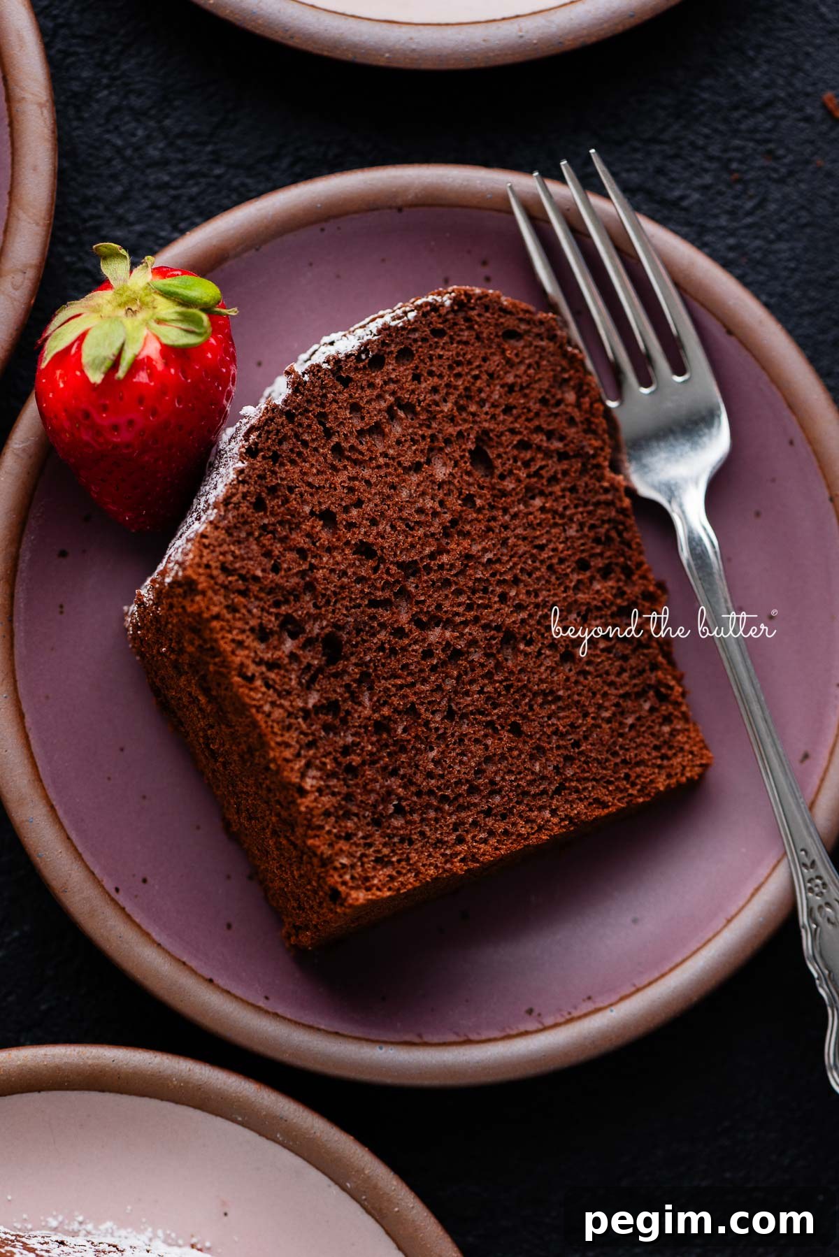Slices of chocolate chiffon cake on small dark purple and light pink ceramic dessert plates with a fork and strawberry placed on each one on a black background.