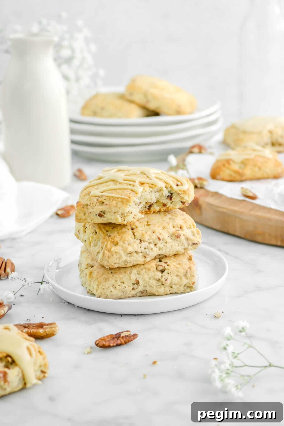 stacked scones on white plate with a bite missing from one with more scones behind, flowers, pecans, and stacked plates