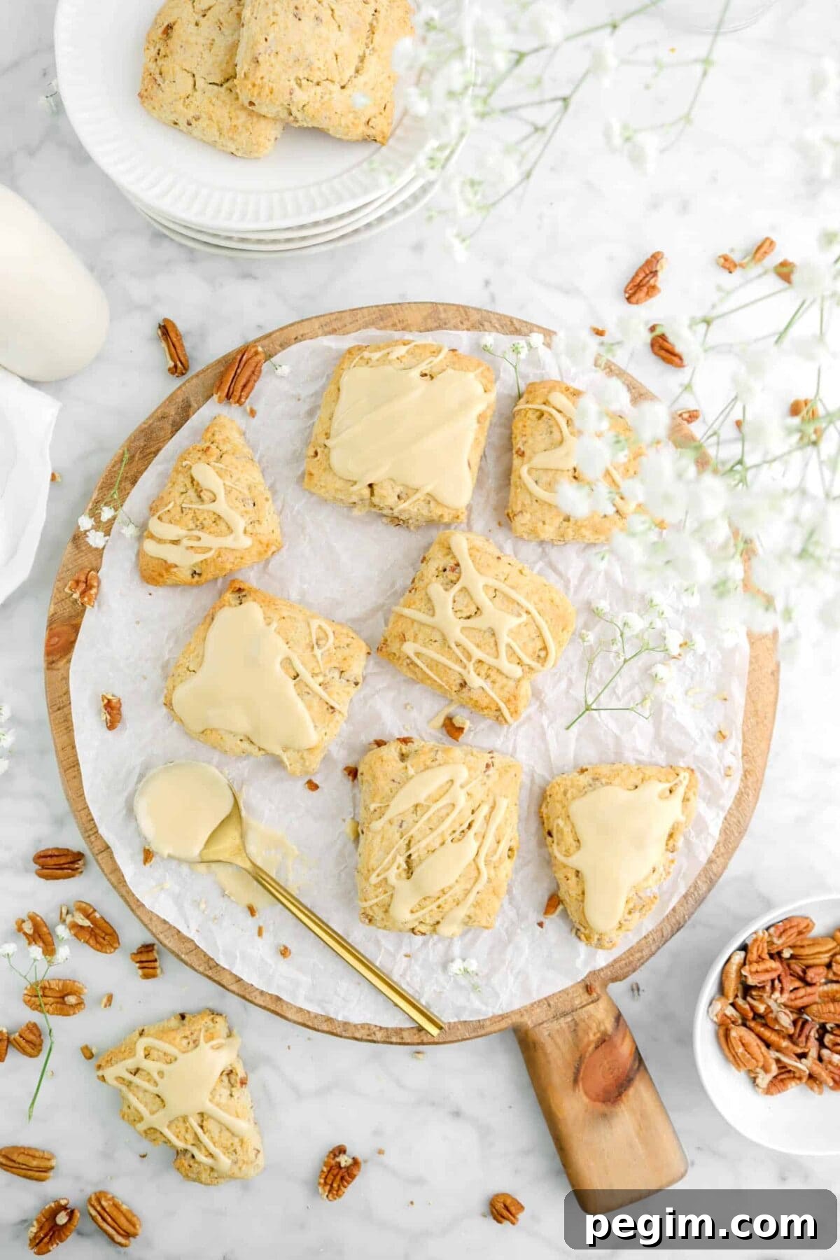 scones on wood serving board with flowers above, a gold spoon with icing and whole pecans scattered around