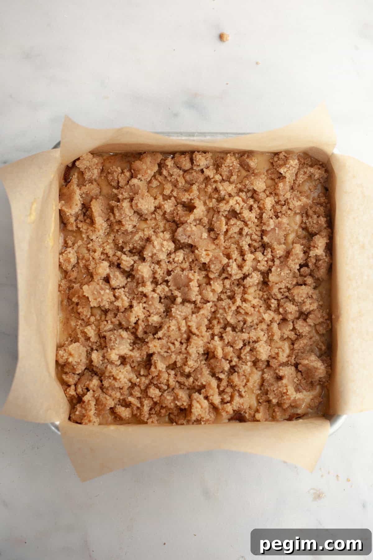 A square baking pan lined with parchment paper and filled with cake batter, topped with streusel.