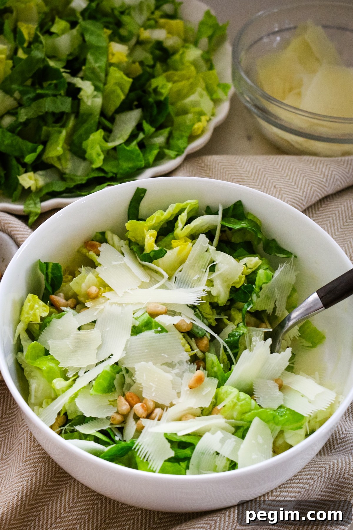 Romaine salad in a white bowl with a fork in it, and a large white plate with romaine salad behind it.