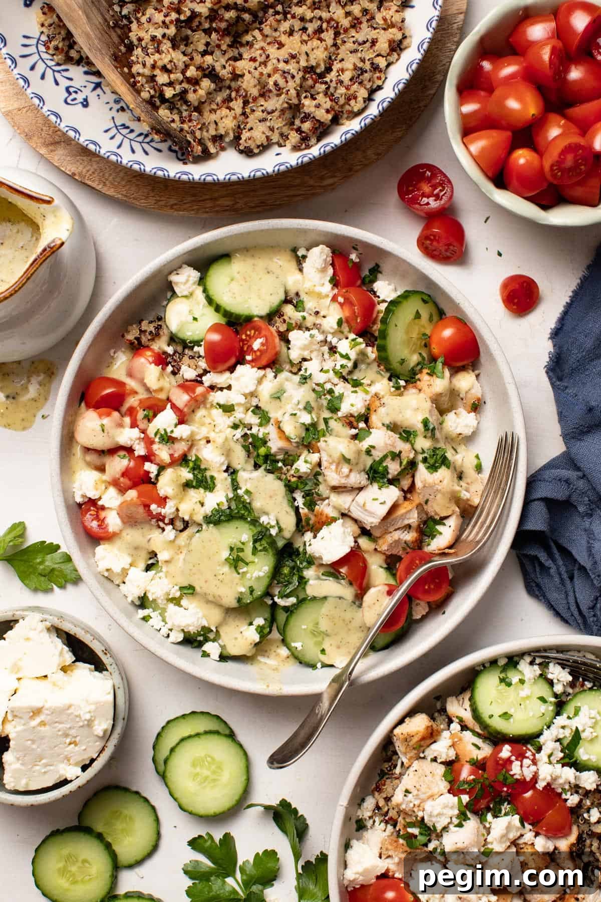 A Mediterranean chicken quinoa bowl next to a bowl with tomatoes and cucumbers, a bowl with feta, and a big bowl of cooked quinoa to the side.
