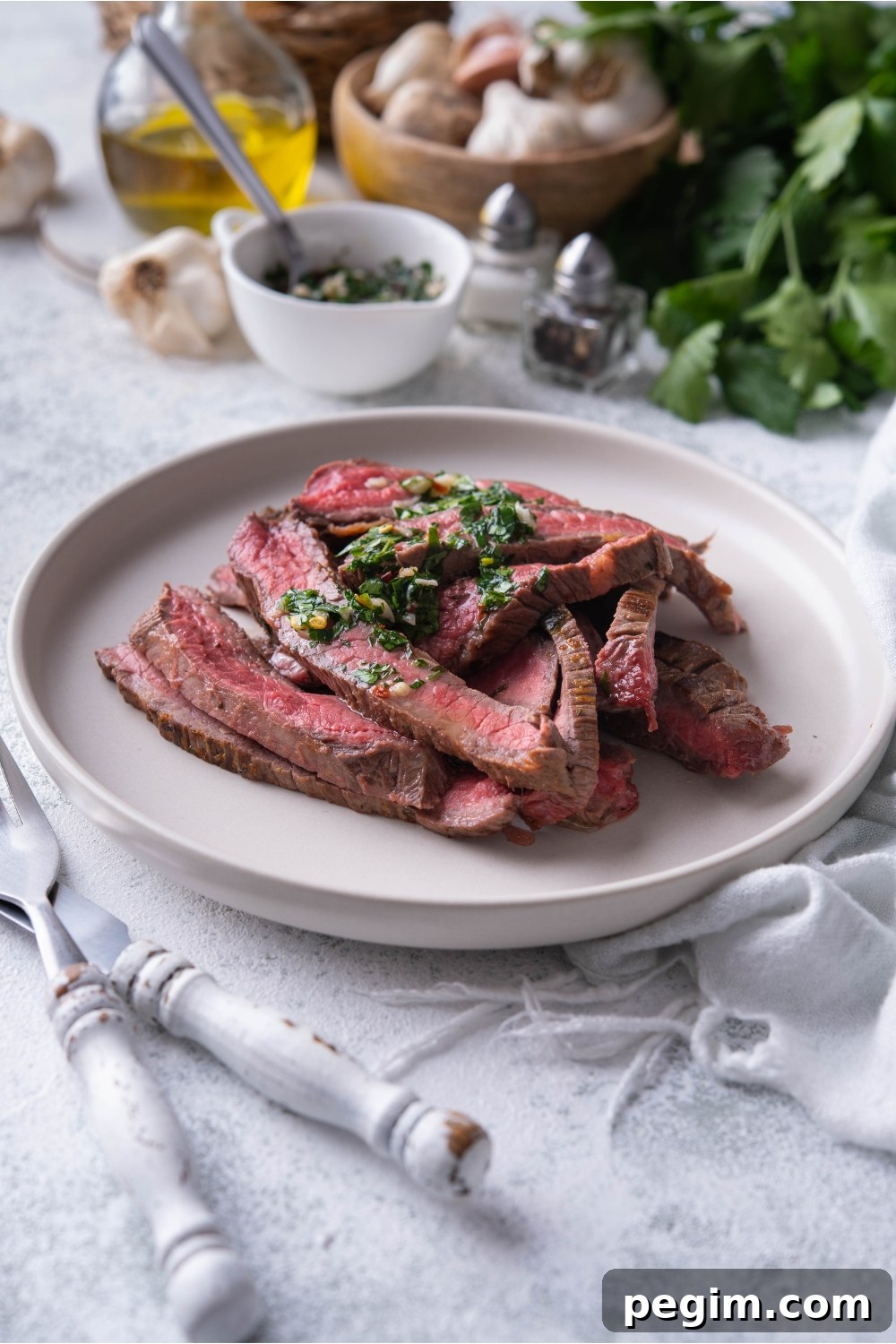 Thinly sliced bavette steak piled on a white plate and covered in chimichurri sauce.